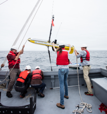 Photo : Cinq personnes a bords une navire, portant des vestes de sauvetage et des casques. Ils ajustent un pièce d’équipement de recherche. 