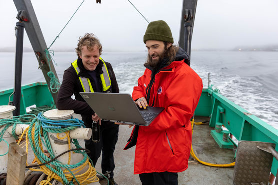 Deux personnes se penchent sur un ordinateur portable sur le pont d’un navire en mer.
