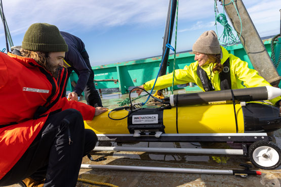 Trois personnes manipulent un véhicule autonome jaune en forme de torpille et mesurant environ 1,5 mètre sur le pont d’un navire en mer.
