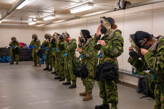 Des membres des Forces armées canadiennes portant une tenue de protection CBRN se tiennent en ligne avec un respirateur sur le visage. Une cagoule est fixée autour du respirateur.