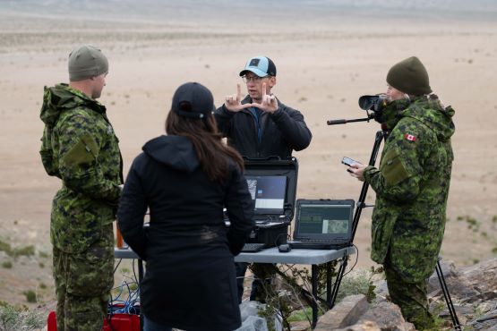 Two civilians and two Canadian Armed Forces members have a conversation around a table stacked with computer screens and technological equipment.