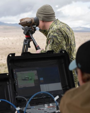 A Canadian Armed Forces member looks through binoculars on a tripod. Another person looks at a computer screen in the foreground