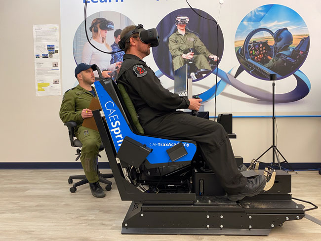 Une personne vêtue d’une combinaison noire utilise un casque de réalité virtuelle dans un siège spécialisé doté d’un gouvernail de direction et de pédales. Une deuxième personne regarde la scène en tenant une planchette à pince.
