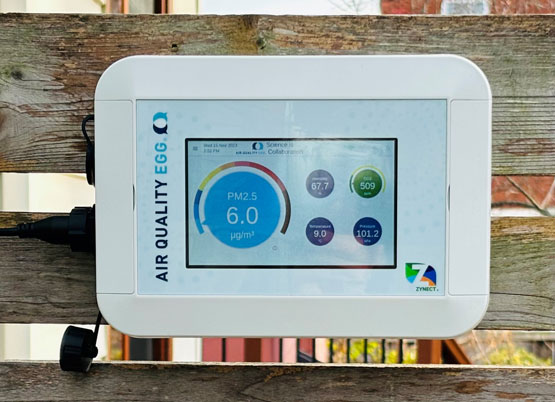 Appareil électronique comprenant un écran qui mesure la qualité de l’air ambiant, accroché à une clôture de bois.