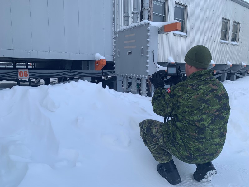 Un membre des Forces armées canadiennes dirige une caméra thermique sur l’extérieur d’un bâtiment; un tas de neige se trouve devant celui-ci.