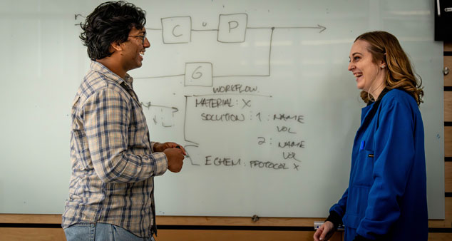 Two scientists conversing in front of a whiteboard with scientific notes