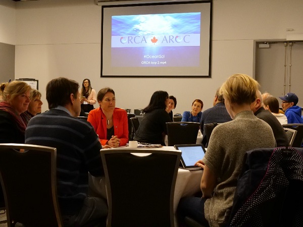 Un auditoire discute, assis autour de tables rondes. Sur le mur en arrière-plan, une projection représente des vagues et le logo (bilingue) de l’AROC.