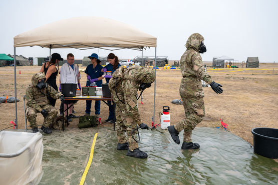 Des soldats en tenue de protection sont debout en rangée devant une table; une équipe se trouvant derrière la table les observe