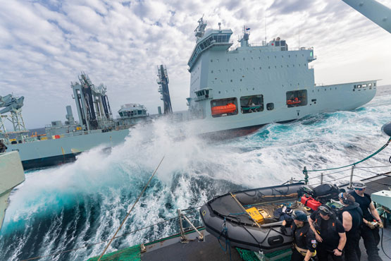 Un grand navire gris vu depuis le pont d’un autre navire. Une grande vague déferle entre les deux navires.