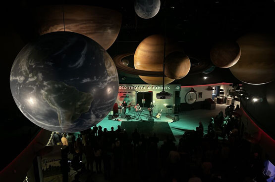 Une salle à l’éclairage tamisé décorée avec des répliques de planètes suspendues au plafond et un groupe de musique jouant sur une scène devant un grand public debout.