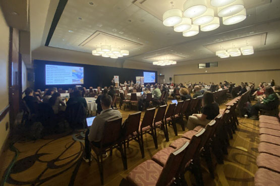 Une grande salle pour la réunion de l’AROC 2023 avec plusieurs participants assis sur des chaises en rangées qui assistent à une présentation avec des diapositives projetées sur deux grands écrans à l’avant de la salle.