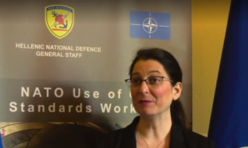 Une femme est devant une pancarte sur laquelle se trouvent le symbole de l’OTAN et le texte suivant : « État-major de la Défense nationale de la Grèce. Atelier de l’OTAN sur l’utilisation des normes civiles. » [(traduction de l’anglais]